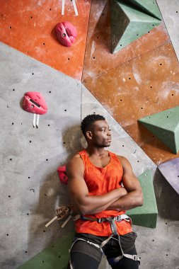 Spor düşkünü Afro-Amerikan bir adam duvara tırmanırken göğsünde çapraz kollar ve kayalarla poz veriyor.