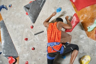 athletic young african american man with alpine harness ascending up bouldering wall, back view clipart