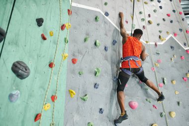 back view of young african american man in orange shirt climbing up bouldering wall with safety rope clipart