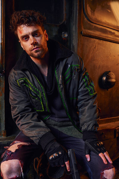 Небритый мужчина в ношеной куртке, держит пистолет и смотрит в камеру в заброшенном постапокалиптическом метро