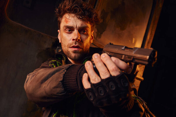 concentrated man with unshaven scratched face looking away and aiming with gun in dark underground