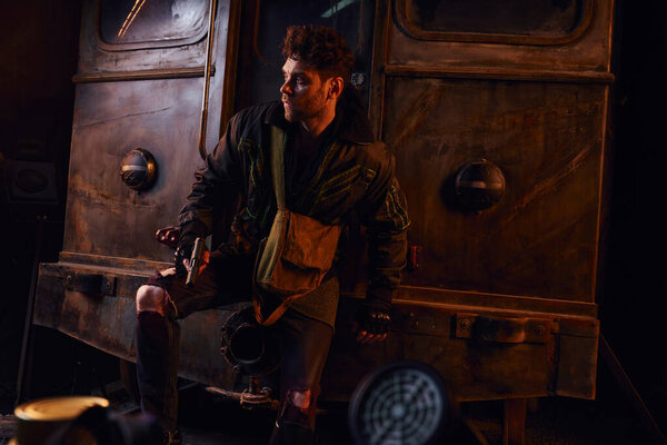 Небритый мужчина в изношенном наряде, стоящий с пистолетом возле ржавой кареты в заброшенном метро после катастрофы