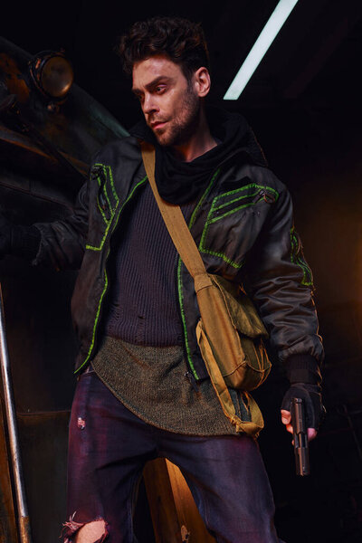 gloomy unshaven man in worn outfit standing with gun and canvas bag in dark abandoned subway