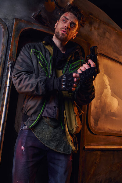 unshaven man in worn clothes and fingerless gloves holding gun and looking away in abandoned subway