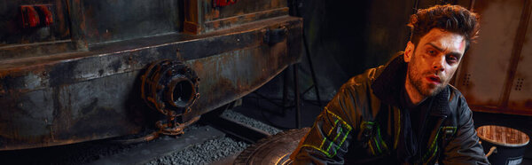 post-apocalyptic survivor with unshaven and injured face near rusty carriage of dark subway, banner
