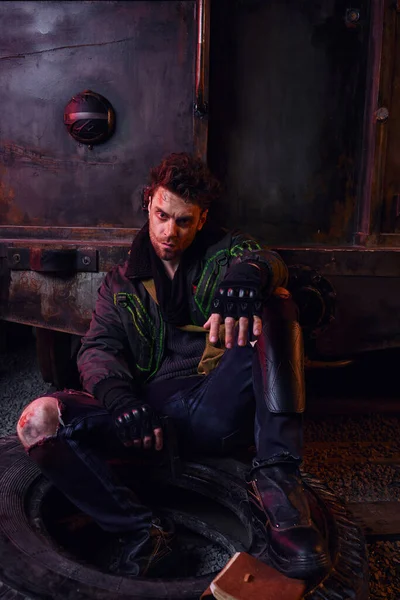 stock image grim man in worn outfit sitting on tire with gun in abandoned subway, life after nuclear catastrophe