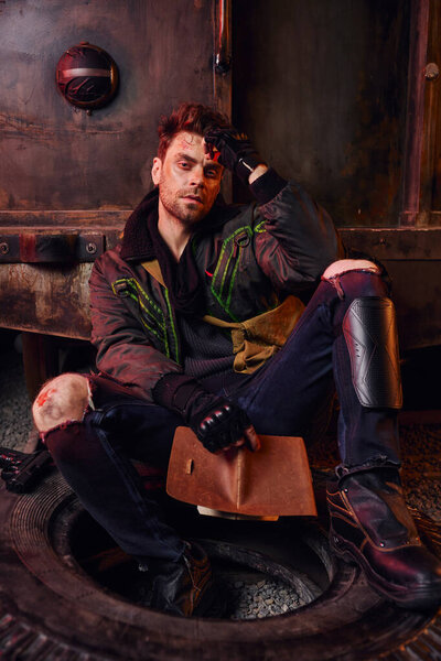 depressed man looking at camera while sitting with diary near rusty carriage in post-disaster subway