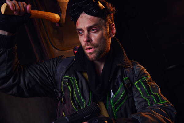 unshaven man in worn clothes with military goggles, axe and gun in dark post-disaster subway