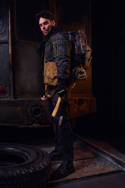 post-apocalyptic survivor with backpack, water bottle and axe looking at camera in abandoned subway