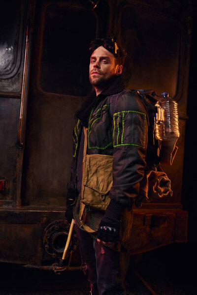 unshaven man with backpack, water bottle and axe looking at camera in dark post-disaster subway