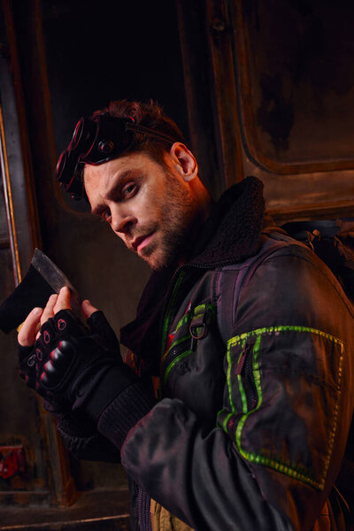 confident man with military goggles and sharp axe looking at camera in post-apocalyptic underground