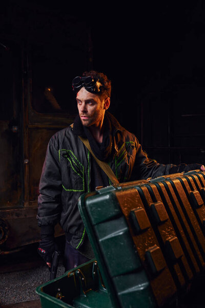 man with military goggles and gun looking away near weapon box in darkness of post-disaster subway