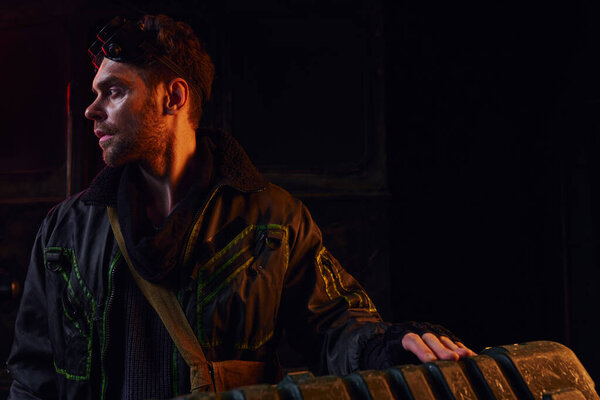 man with military goggles looking away near weapon box in red light of post-apocalyptic subway