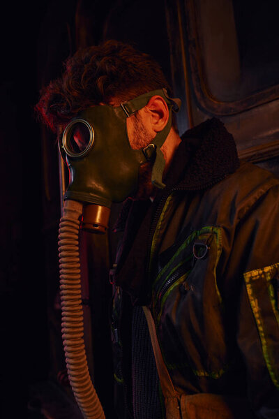 man in mask and worn jacket in red light of dark underground tunnel, post-apocalyptic survival