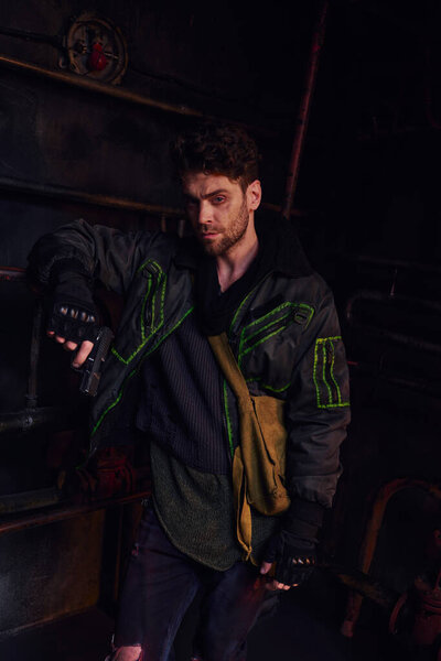 вдумчивый человек в грязной одежде, стоящий с пистолетом возле ржавой кареты в метро после катастрофы