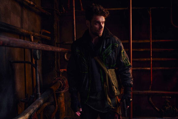 серьезный человек в изношенном наряде, смотрящий в сторону ржавых трубопроводов заброшенного метро, постапокалипсиса