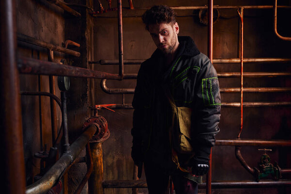 безнадежный человек в грязной одежде возле ржавых трубопроводов в подземном туннеле, постапокалиптическая концепция