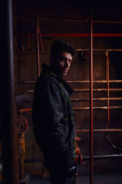 despaired unshaven man looking at camera next to rusty pipelines in abandoned post-disaster subway