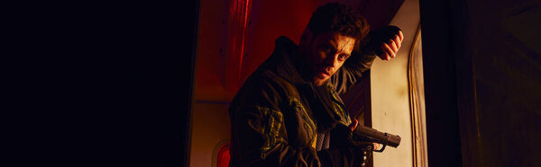 unshaven man with gun looking at camera in abandoned subway carriage with red light, banner