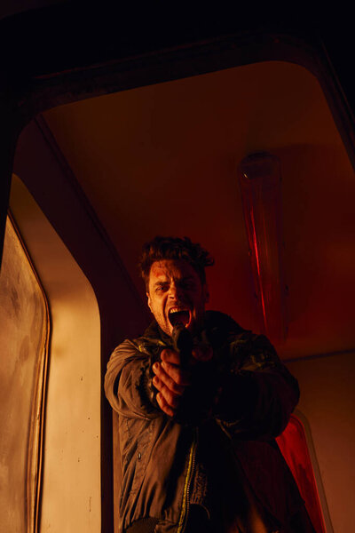 screaming man looking at camera and aiming with gun in red light of abandoned subway carriage