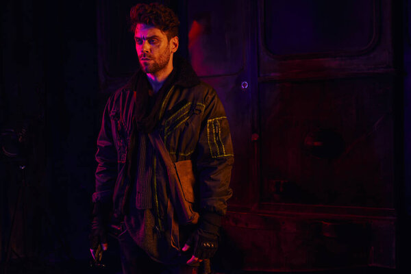 отчаявшийся небритый мужчина в изношенной одежде, стоящий в темном постапокалиптическом метро с красным светом