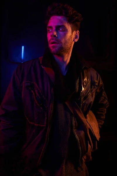 tense man in black jacket looking away in darkness of abandoned underground tunnel with neon light