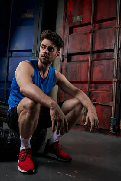 Spor giyimli atletik bir adam konteynırların yanında oturuyor ve dışarı bakıyor.