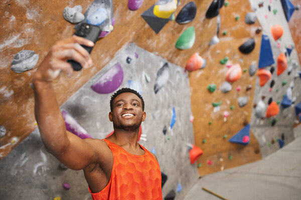 happy african american man taking selfie and smiling cheerfully with climbing wall backdrop