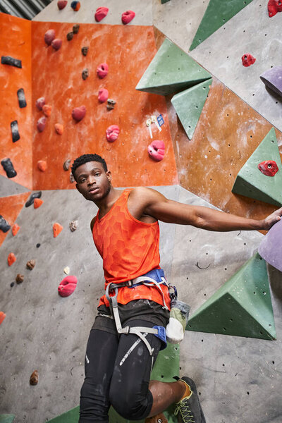 handsome african american man with alpine harness in orange shirt posing next to rock climbing wall