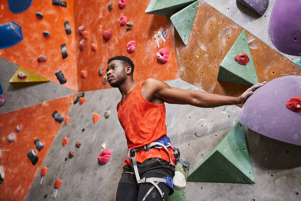 good looking african american man with alpine harness posing near climbing wall and looking away