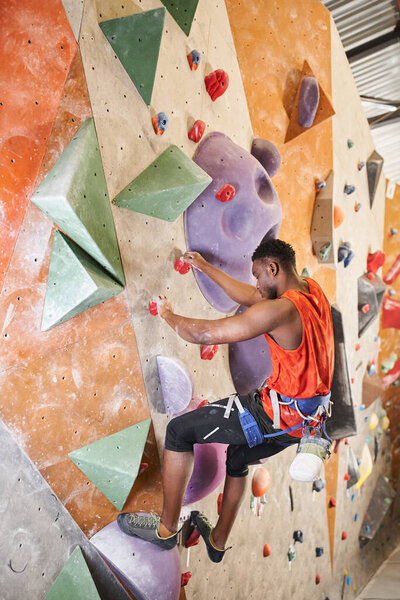 vertical shot of sporty african american man with alpine harness climbing up rock wall, bouldering