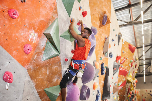 handsome african american man climbing up the wall with alpine harness and looking at camera
