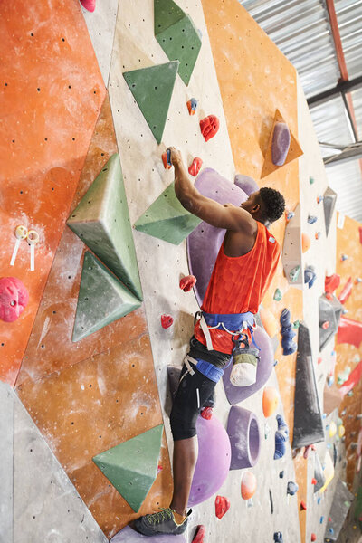 vertical shot of african american man with alpine harness in orange shirt climbing up the wall