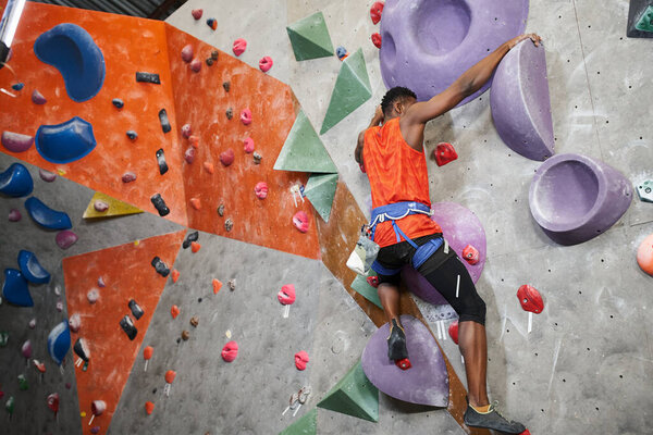 sporty african american man in orange shirt with alpine harness climbing up wall, sport concept