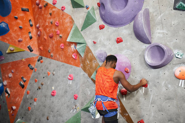 strong african american man in orange shirt climbing up rock wall with alpine harness, bouldering
