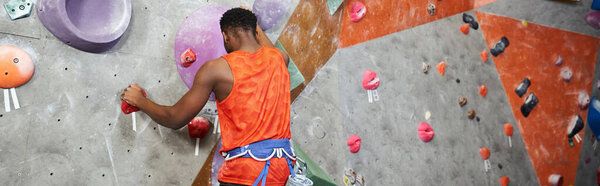 sporty african american man in orange shirt climbing up rock wall with alpine harness, banner