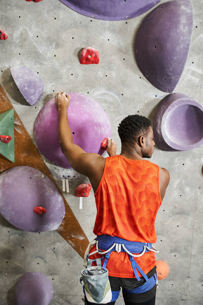 vertical shot of muscular african american man in orange shirt climbing up rock wall, bouldering