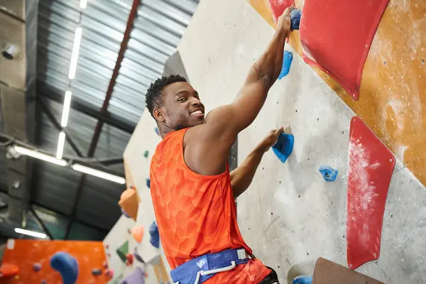 stock image cheerful african american man smiling happily at camera while climbing up rock wall, bouldering