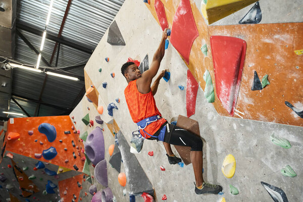 athletic african american man posing in profile with alpine harness climbing and looking at camera