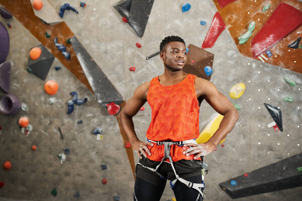 athletic african american man in orange shirt with alpine harness posing with hands on hips