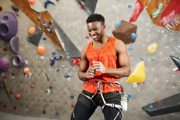 cheerful african american man in orange shirt laughing sincerely posing next to rock climbing wall