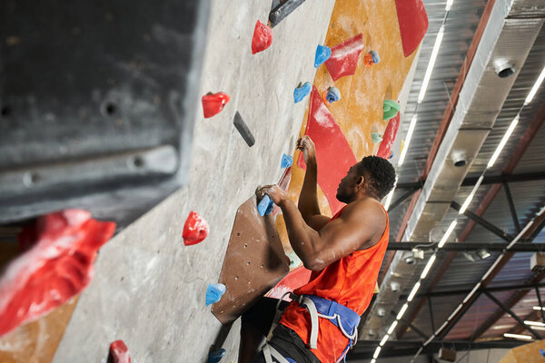 handsome african american man in orange shirt with alpine harness climbing up bouldering wall