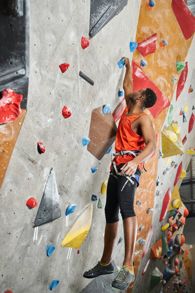 vertical shot of young african american man hanging on his fingertips on bouldering rock wall