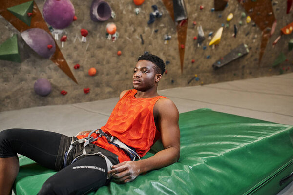 young african american man with alpine harness chilling on crash pad after climbing up rock wall