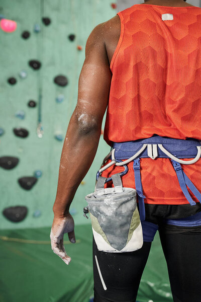 cropped view of african american man posing next to climbing rock wall with alpine harness