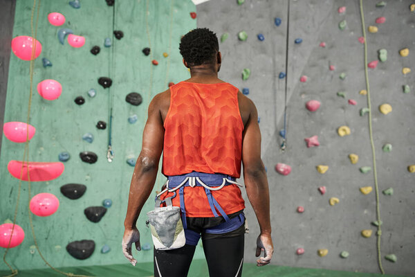 back view of young african american man in orange shirt with alpine harness near bouldering wall