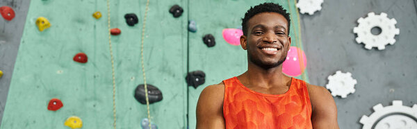 joyous african american man in orange shirt posing near bouldering wall smiling at camera, banner