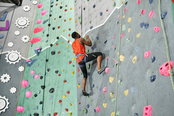 young african american man in orange shirt climbing up rock wall with rope and alpine harness