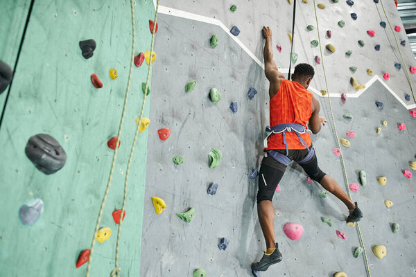 back view of young african american man in orange shirt climbing up bouldering wall with safety rope