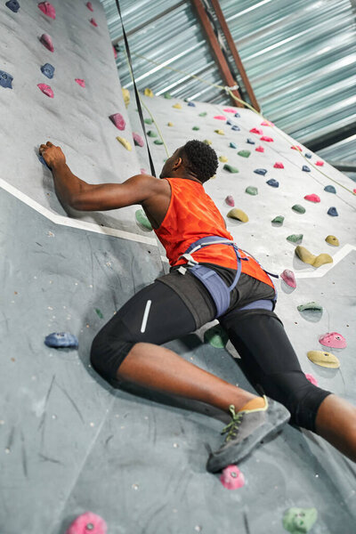 sporty african american man in orange shirt with alpine harness ascending up rock wall, safety rope